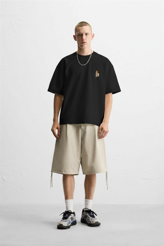 Person wearing a black t-shirt with a small logo and beige shorts on a white background